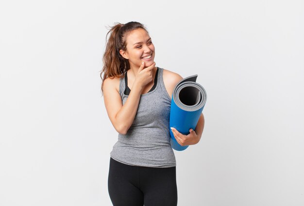 Jeune jolie femme souriante avec une expression heureuse et confiante avec la main sur le menton et tenant un tapis de yoga