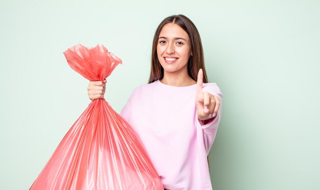 Jeune jolie femme souriante fièrement et avec confiance faisant numéro un. notion de poubelle