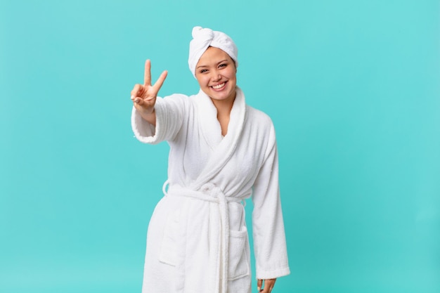 Jeune jolie femme souriante et heureuse, faisant des gestes de victoire ou de paix et portant un peignoir après la douche