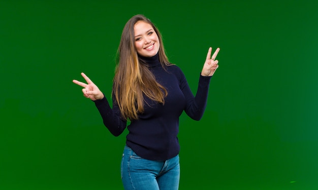 Jeune jolie femme souriante et à la joyeuse, sympathique et satisfaite, gesticulant la victoire ou la paix à deux mains contre le mur vert