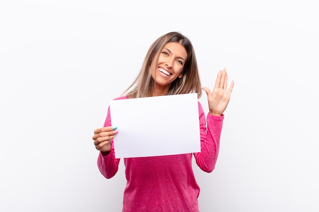 Jeune jolie femme souriante joyeusement et joyeusement, agitant la main, vous accueillant et vous saluant, ou disant au revoir tenant une pancarte