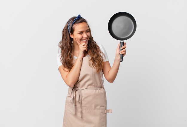 Jeune jolie femme souriante joyeusement et rêvant ou doutant du concept de chef et tenant une casserole