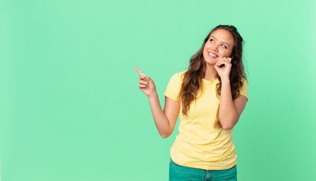 Jeune jolie femme souriante joyeusement et rêvant ou doutant et pointant vers l'espace de copie