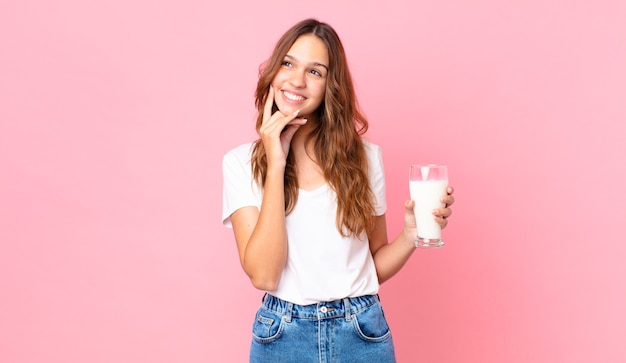 Jeune jolie femme souriante joyeusement et rêvant ou doutant et tenant un verre de lait