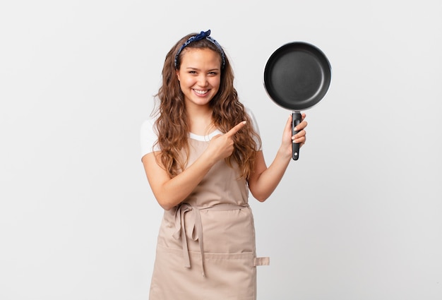 Jeune jolie femme souriante joyeusement, se sentant heureuse et pointant vers le concept de chef latéral et tenant une casserole