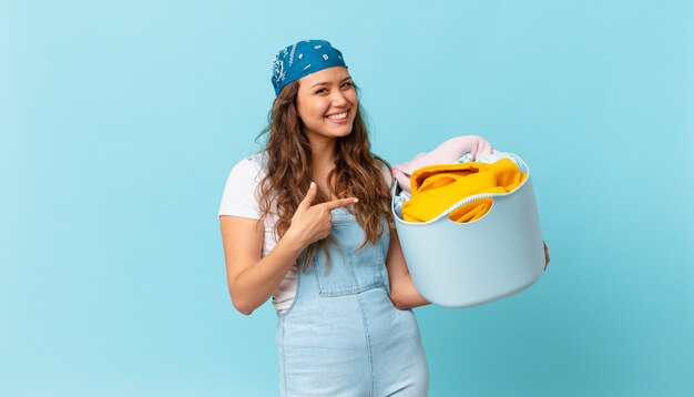 Jeune jolie femme souriante joyeusement, se sentant heureuse et pointant vers le côté et tenant un panier à linge