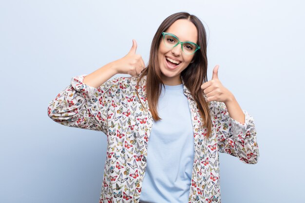 Jeune jolie femme souriante largement à la recherche de bonheur, positive, confiante et réussie, avec les deux pouces contre le mur bleu