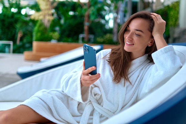 Jeune jolie femme souriante portant un peignoir blanc à l'aide de smartphone pour regarder la vidéo et la navigation en ligne en position couchée sur une chaise longue pendant la détente au spa