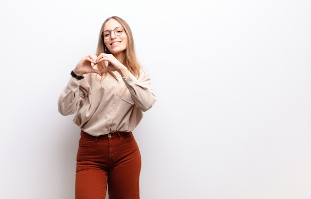 Jeune jolie femme souriante et se sentant heureuse, mignonne, romantique et amoureuse, en forme de cœur avec les deux mains contre le mur blanc