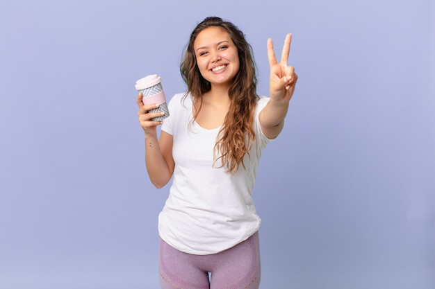 Jeune jolie femme souriante et semblant heureuse, gesticulant la victoire ou la paix et tenant un café