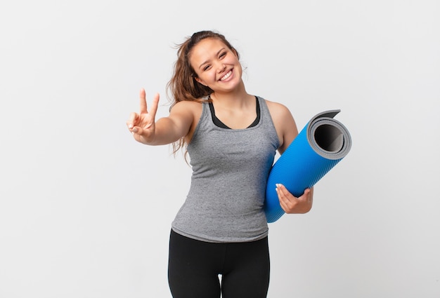 Jeune jolie femme souriante et semblant heureuse, gesticulant la victoire ou la paix et tenant un tapis de yoga