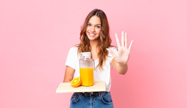 Jeune jolie femme souriante et sympathique, montrant le numéro cinq et tenant un plateau avec un jus d'orange