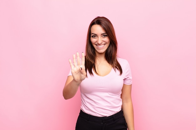 Jeune jolie femme souriante et sympathique, montrant le numéro quatre ou quatrième avec la main vers l'avant, compte à rebours contre le mur rose