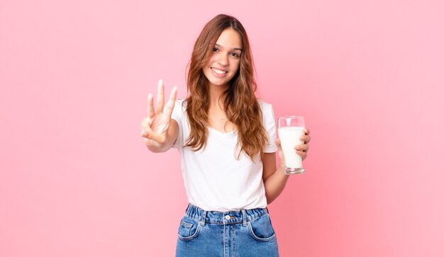 Jeune jolie femme souriante et sympathique, montrant le numéro trois et tenant un verre de lait