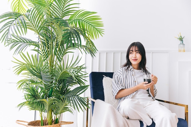 Jeune jolie femme sourit et s'assoit sur la chaise en se relaxant en buvant une tasse de café chaud dans le salon moderne avec une plante verte une réflexion de guérison dans un environnement intérieur dans une maison confortable