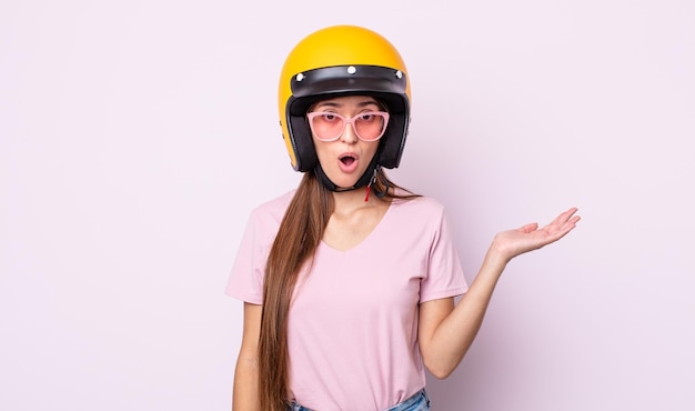 Jeune jolie femme à la surprise et choquée avec la mâchoire tombée tenant un motard et un casque d'objet