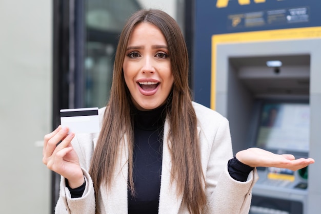 Jeune jolie femme tenant une carte de crédit à l'extérieur avec une expression faciale choquée