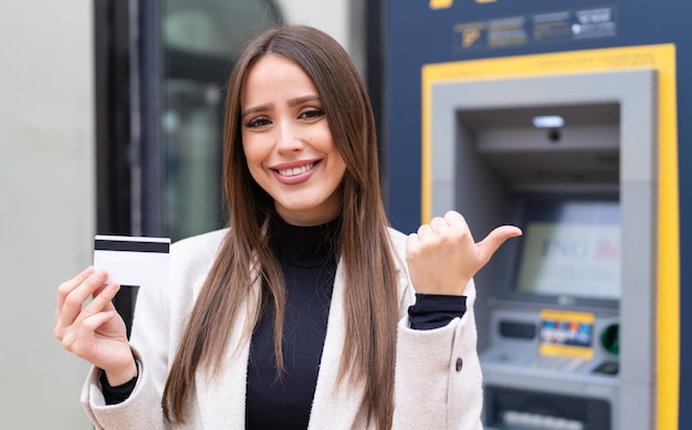 Jeune jolie femme tenant une carte de crédit à l'extérieur pointant vers le côté pour présenter un produit