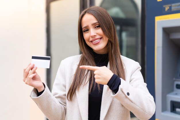 Jeune jolie femme tenant une carte de crédit à l'extérieur et la pointant