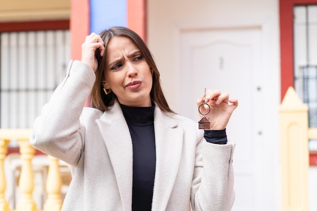 Jeune jolie femme tenant les clés de la maison à l'extérieur ayant des doutes et avec une expression de visage confuse