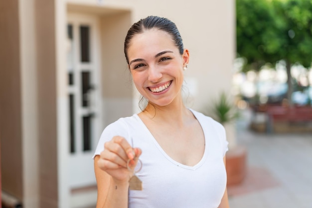 Jeune jolie femme tenant les clés de la maison à l'extérieur souriant beaucoup