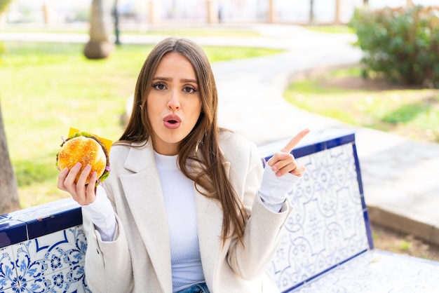 Jeune jolie femme tenant un hamburger à l'extérieur surpris et pointant du côté