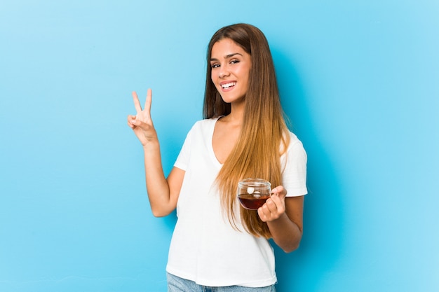 Jeune jolie femme tenant une tasse de thé joyeuse et insouciante montrant un symbole de paix avec les doigts