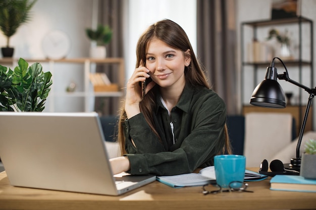 Jeune jolie femme en tenue décontractée parlant sur téléphone portable et tapant sur ordinateur portable