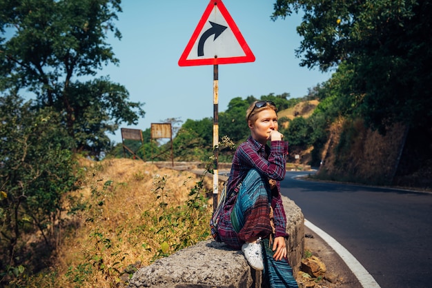 Photo jeune jolie femme touriste sur le bord de la route en attente d'une voiture qui passe sur une journée ensoleillée. blonde élégante faisant de l'auto-stop en asie. liberté, indépendance, aventure