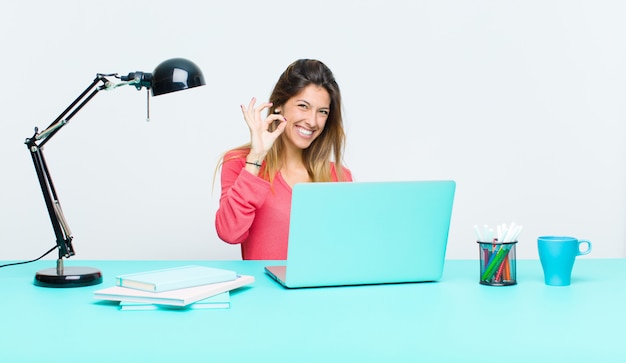 Jeune jolie femme travaillant avec un ordinateur portable se sentant heureuse, détendue et satisfaite, montrant son approbation avec un geste correct, souriant