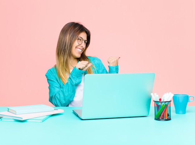 Jeune jolie femme travaillant avec un ordinateur portable souriant gaiement et avec désinvolture, pointant du côté de la surface, se sentant heureuse et satisfaite