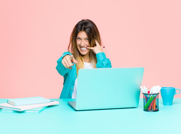 Jeune jolie femme travaillant avec un ordinateur portable souriant joyeusement et pointant vers la caméra tout en faisant un appel, vous gestes plus tard, parler au téléphone