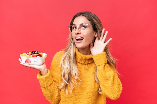 Jeune jolie femme uruguayenne tenant un bol de fruits isolé sur fond rouge écoutant quelque chose en mettant la main sur l'oreille