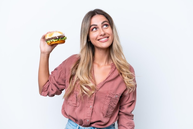 Jeune jolie femme uruguayenne tenant un hamburger isolé sur fond blanc levant les yeux en souriant