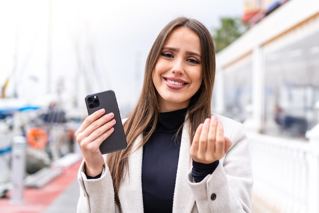 Jeune jolie femme utilisant un téléphone portable à l'extérieur invitant à venir avec la main Heureux que vous soyez venu