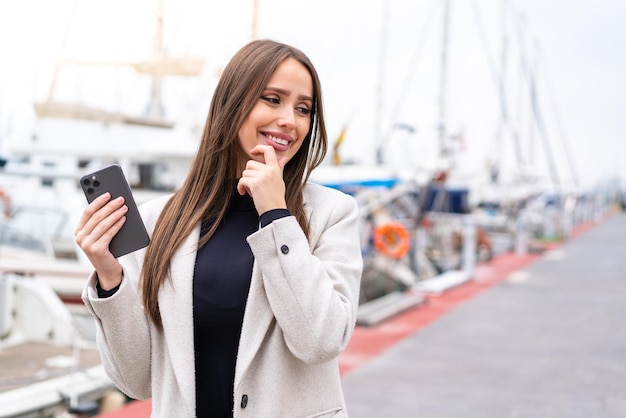 Jeune jolie femme utilisant un téléphone portable à l'extérieur en pensant à une idée et en regardant de côté