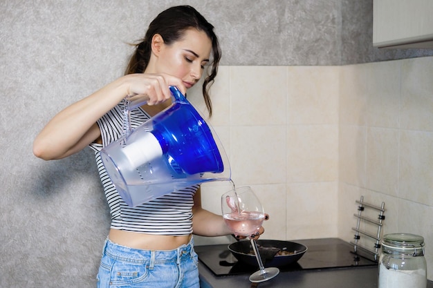 Une jeune jolie femme verse de l'eau filtrée propre à partir d'une cruche en plastique dans la cuisine