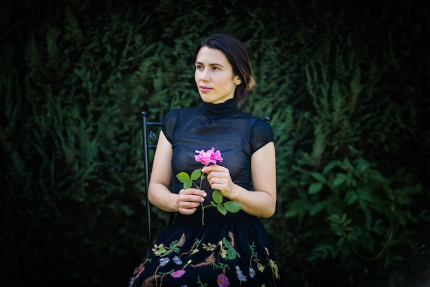 Jeune jolie femme en vêtements noirs est assise sur une chaise dans le jardin près d'un buisson avec une rose dans les mains