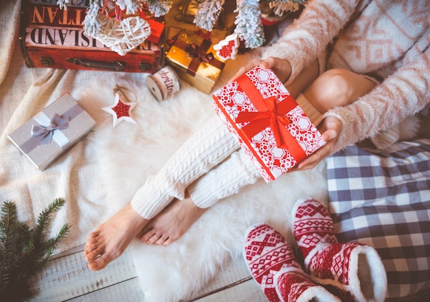 Une Jeune Jolie Femme Vêtue De Vêtements Chauds Et Confortables Est Assise Sur Le Parquet Clair Dans Sa Maison Lumineuse Près De L'arbre De Noël Et Tient Une Boîte-cadeau Dans Ses Mains.