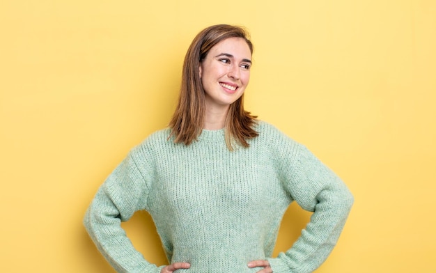 Jeune jolie fille à l'air heureuse, gaie et confiante, souriante fièrement et regardant de côté avec les deux mains sur les hanches