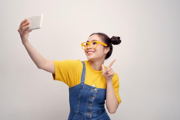 Jeune jolie fille asiatique qui porte une salopette en jean et des lunettes jaunes prenant un selfie en souriant