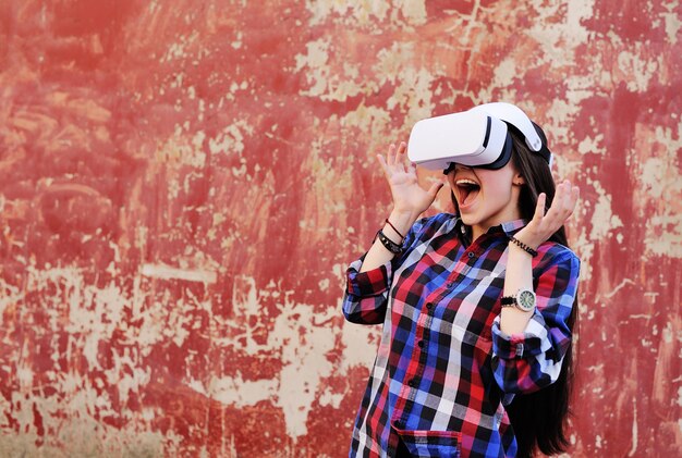 Jeune jolie fille aux cheveux longs dans des verres VR en chemise à carreaux sur la surface du mur rouge grunge.