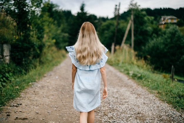 Jeune jolie fille blonde élégante en robe bleue posant de retour sur la route à la campagne