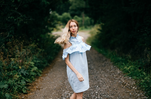 Jeune jolie fille blonde élégante en robe bleue posant sur la route à la campagne