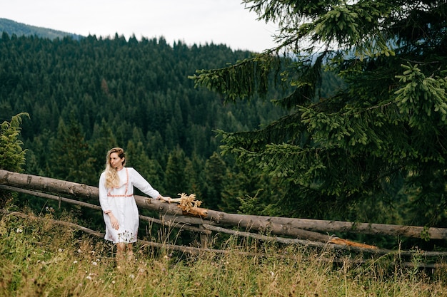 Jeune jolie fille blonde en robe blanche avec broderie posant avec bouquet d'épillets sur paysage pittoresque