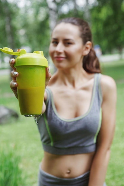 Une jeune jolie fille boit une boisson énergisante dans un verre en plastique tout en faisant du jogging dans un parc d'été verdoyant