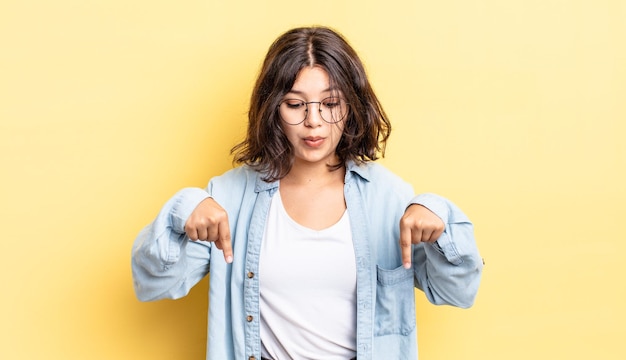 Jeune jolie fille avec la bouche ouverte pointant vers le bas avec les deux mains, l'air choquée, étonnée et surprise