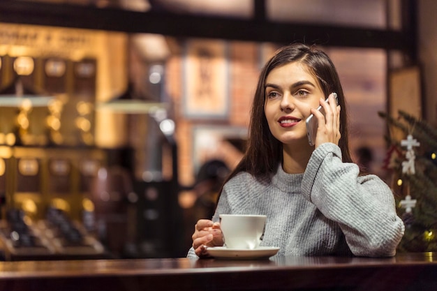 Une jeune jolie fille brune porte un pull gris a une conversation agréable par smartphone assis près de la fenêtre du café