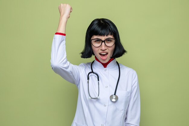 Jeune jolie fille caucasienne agacée avec des lunettes optiques en uniforme de médecin avec stéthoscope levant le poing