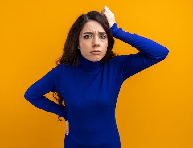 Jeune jolie fille confuse gardant la main sur la taille et sur la tête en regardant la caméra